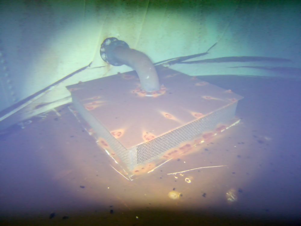 A mesh strainer box on the floor of a steel water tank, surrounded by settled sediment.